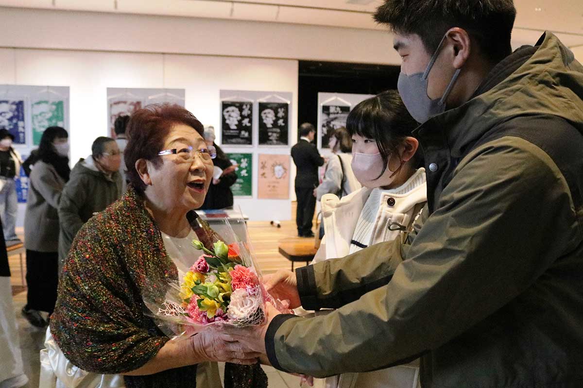 「おつかれさま！」お孫さんから花束を贈られ、喜びの笑顔を輝かせる土橋郁子さん（左）