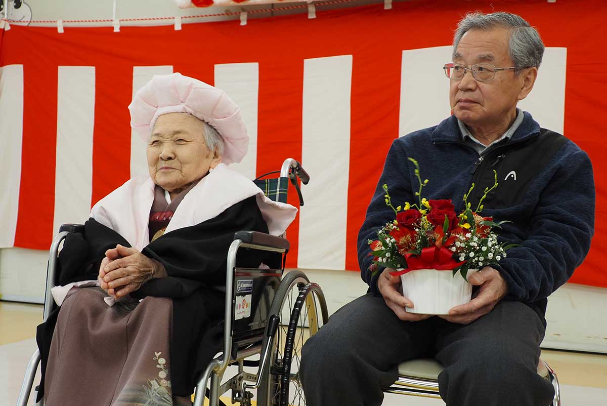 桃色のちゃんちゃんこと帽子を身に着けた100歳の盛トセさん（左）、長男の久男さん
