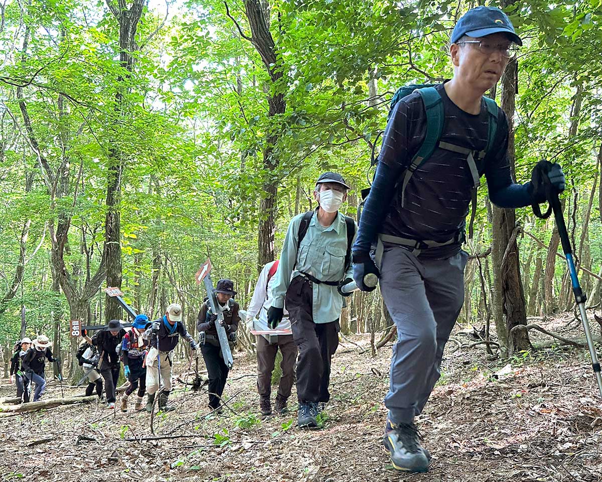 合目標識や穴を掘るための道具などを持ちながら山道を歩く