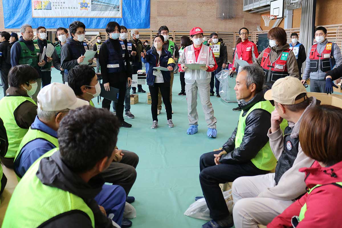 外部から入った保健医療福祉の支援者らが釜石市の担当者から避難所の状況の説明を受ける=県総合防災訓練・釜石東中会場