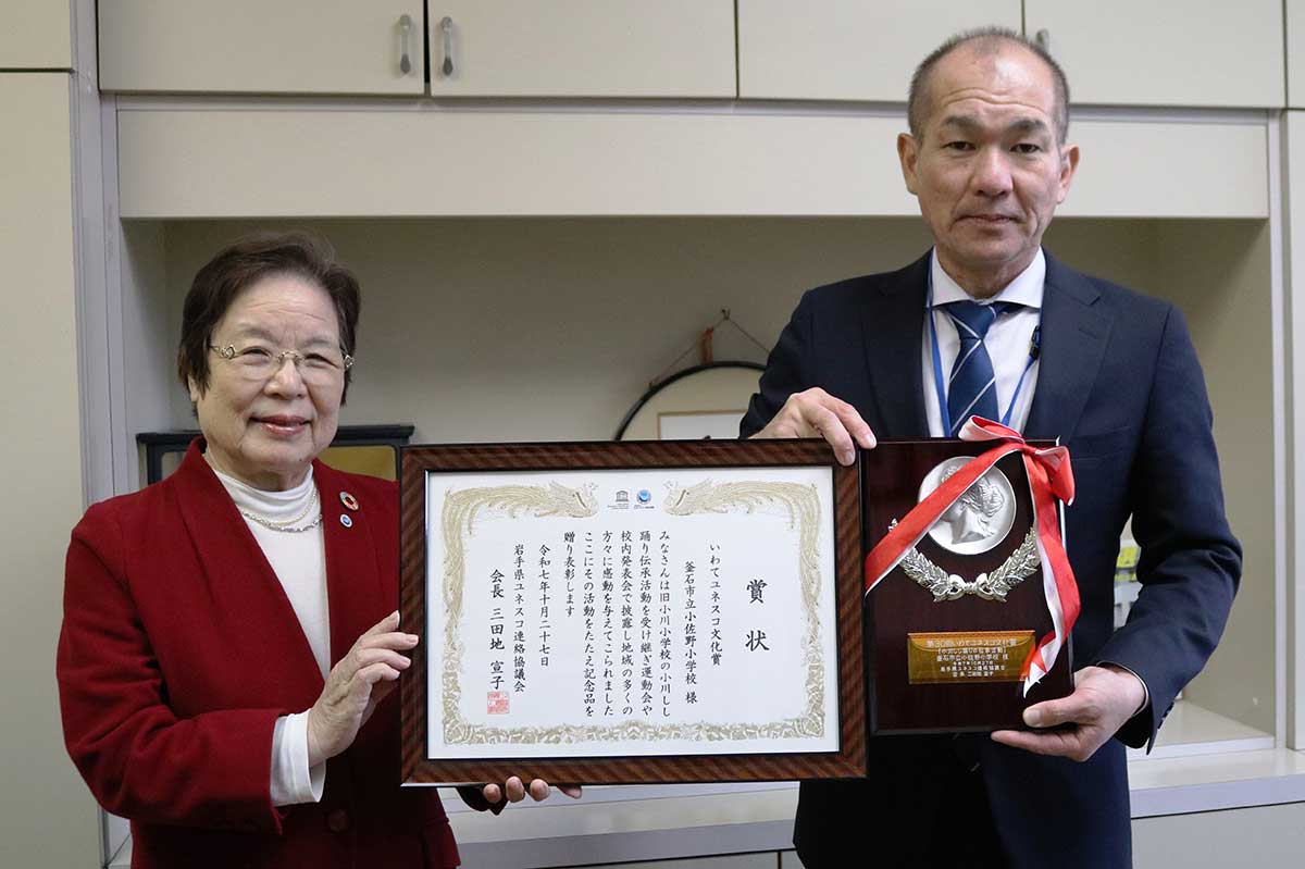 県ユネスコ連絡協に代わり、釜石ユネスコ協会の岩切久仁会長（左）が小佐野小の松本孝嗣校長（右）に表彰状と盾を届けた＝11月26日、同校