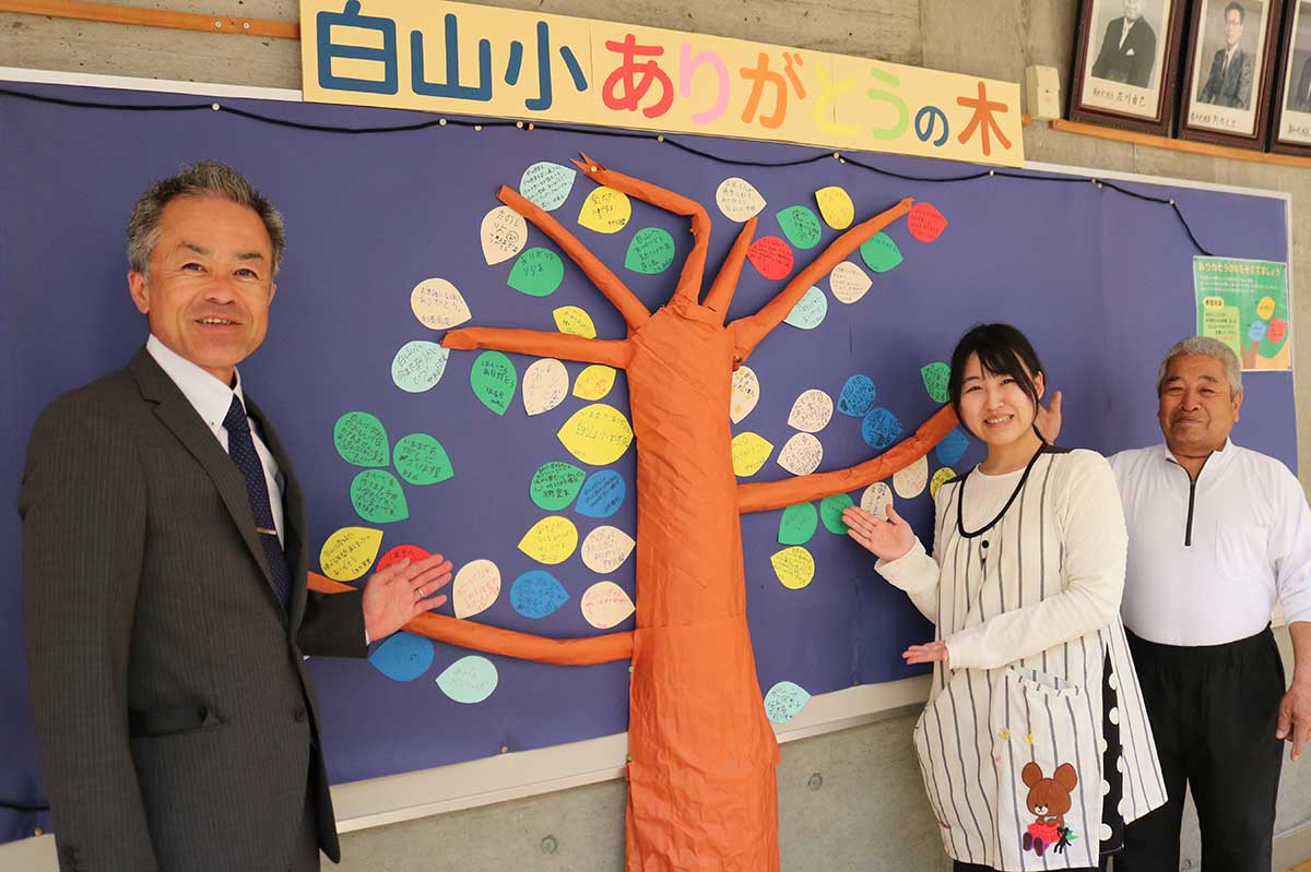 来校者のメッセージを掲示する「ありがとうの木」。鈴木慎校長（左）らがPR。年度末には白山小への思いが“大樹”となる