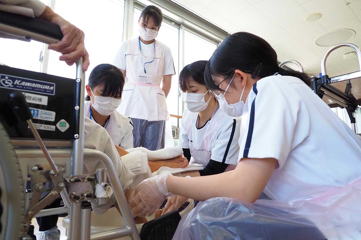 先輩看護師に教わりながら足を拭く生徒たち